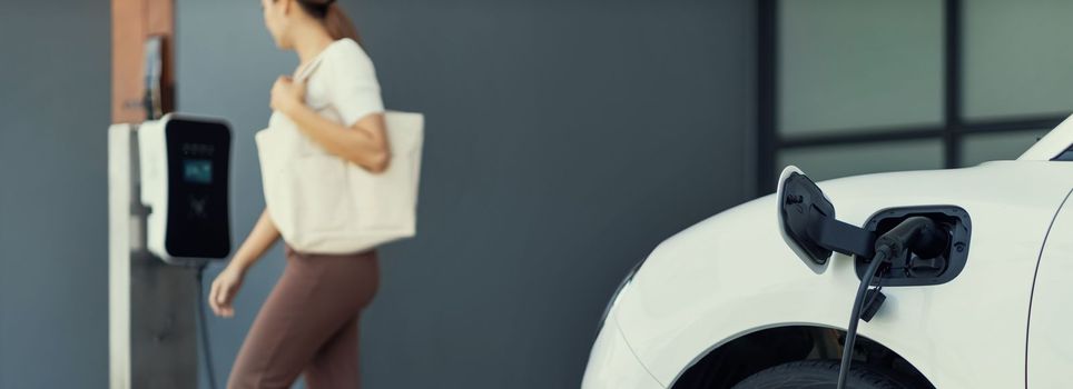 Focus image of electric vehicle recharging battery at home charging station with blurred woman walking in the background. Progressive concept of green energy technology applied in daily lifestyle.