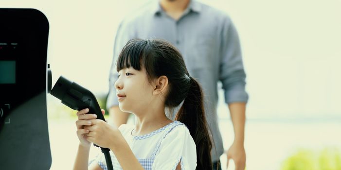 Focus progressive young girl holding EV charger device from home electric charging station for renewable energy powered car with blur asian man background. Sustainable green energy technology concept.