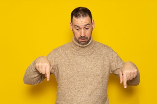Bearded handsome latino man wearing a beige turtleneck pointing down with fingers showing advertisement isolated over yellow background