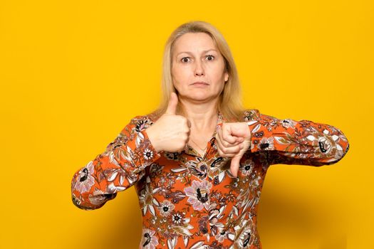 Pretty caucasian blonde woman in a patterned dress giving one thumbs up and one thumbs down isolated over yellow background. doubt concept
