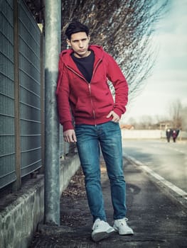 Attractive young man in city street leaning against pole, looking at camera, wearing hoodie sweater and jeans