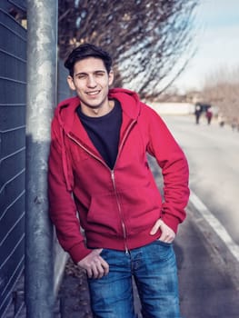 Attractive young man in city street smiling and leaning against pole, looking at camera, wearing hoodie sweater and jeans