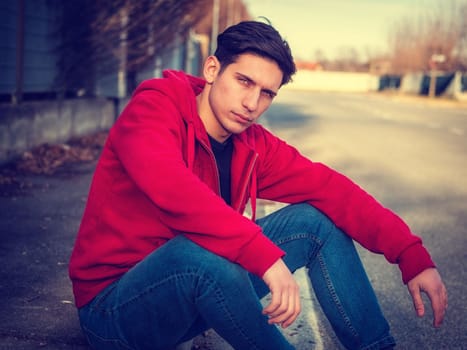 Attractive young man sitting on street curb, looking at camera, alone, wearing hoodie and jeans