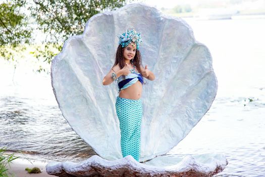 A cute little girl in a mermaid costume stands outdoor, in a large sea shell, holds her thumbs up. Copy space