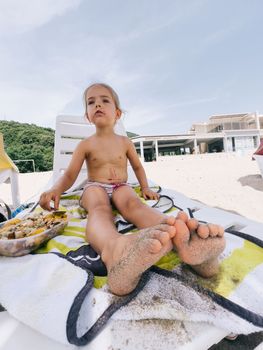 Little girl is sitting on a sun lounger on the beach next to a plastic box with food. High quality photo