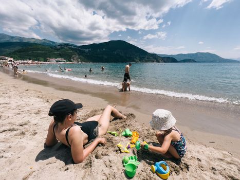 Mom is sunbathing on the beach next to a little girl playing with a toy shovel in the sand. High quality photo
