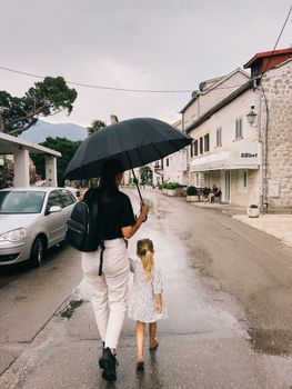 Mom and a little girl are walking down the street under an umbrella holding hands. High quality photo