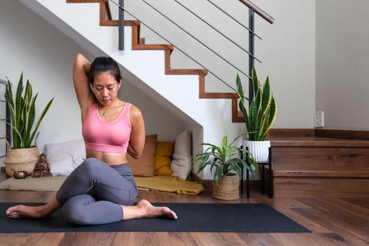 Young Asian woman doing yoga at home stretching arms, shoulders and hips in gomukhasana pose. Cow face asana. Healthy lifestyle concept.