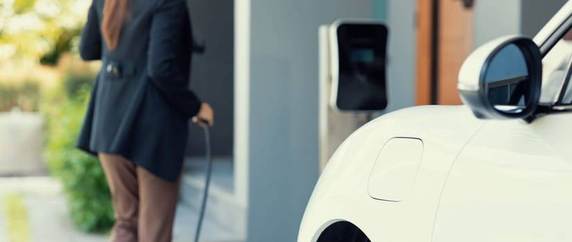 Closeup progressive woman install cable plug to her electric car with home charging station. Concept of the use of electric vehicles in a progressive lifestyle contributes to clean environment.