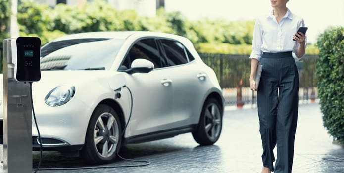 Closeup businesswoman using tablet, walking while recharging her electric vehicle with charging station at public car parking. Progressive lifestyle of technology and ecological concern by EV car