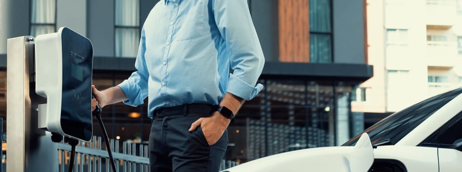 Closeup progressive man holding EV charger plug from public charging station for electric vehicle with background of residential building as concept eco-friendly sustainability energy car concept.