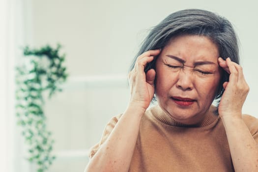 An agonizing senior woman with a headache, compress her temple with both hands at her contented living room. Senior health, nursing home, caretaker service.