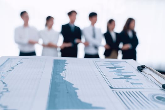 Closeup focus on financial report, performance result papers in front of camera with blur background of office workers, colleagues holding each other hands in line as concept of harmony in workplace.