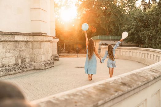 Daughter mother run holding hands. In blue dresses with flowing long hair, they hold balloons in their hands against the backdrop of a sunset and a white building