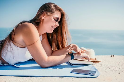 Successful business woman in yellow hat working on laptop by the sea. Pretty lady typing on computer at summer day outdoors. Freelance, travel and holidays concept.