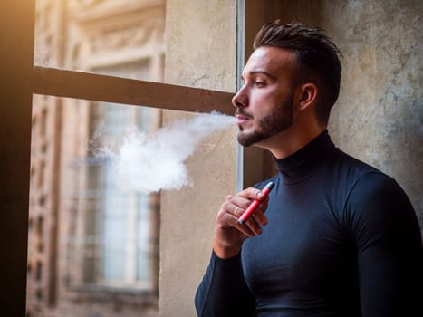 Handsome man vaping, smoking e-cigarette by a window, looking out
