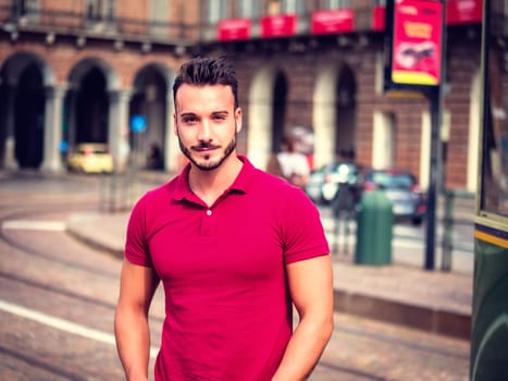 One handsome young man in urban setting in European city, standing, wearing red polo shirt looking at camera