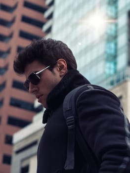 Stylish Young Handsome Man in Black Coat Standing in City Center Street with Skyscraper Behind Him, Looking Down to the Left of the Frame.