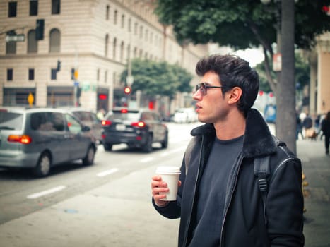 Attractive young man in modern city center drinking coffee from paper cup, wearing leather jacket