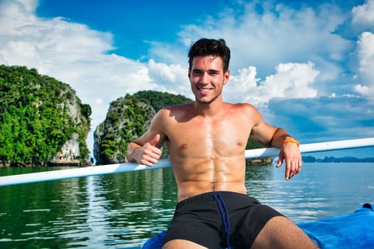 Cheerful young shirtless male with muscular torso smiling and showing thumb up, while leaning against railing of boat floating in sea water near island in sunny summer day