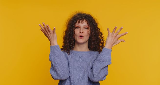 Oh my God, Wow. Excited amazed winner woman raising hands in surprise looking at camera with big eyes, shocked by sudden victory. Sincere young curly haired girl isolated on yellow studio background