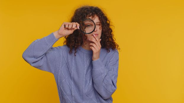 Investigator researcher scientist woman holding magnifying glass near face, looking into camera with big zoomed funny eyes, searching, analysing. Young curly haired girl on yellow studio background