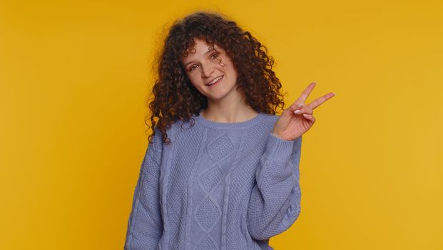 Happy curly haired young woman showing victory v sign, hoping for success and win, doing peace gesture, smiling with kind optimistic expression. Teen girl isolated alone on yellow studio background