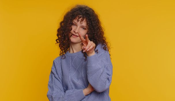 I am watching you. Caucasian woman pointing at her eyes and camera, show I am watching you gesture, spying on someone. Young adult curly haired girl isolated alone on yellow studio wall background