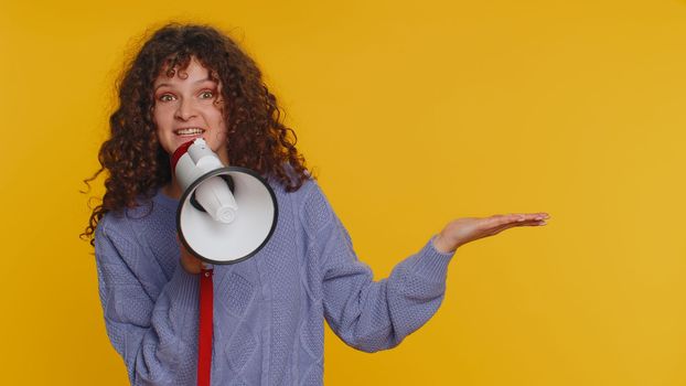 Happy woman talking with megaphone, proclaiming news, loudly announcing advertisement, warning using loudspeaker to shout speech, pointing empty place. Copy-space. Young girl on yellow background