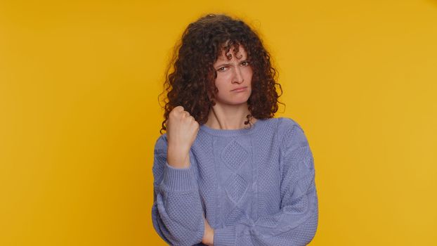 Aggressive angry curly haired woman trying to fight at camera, shaking fist, boxing with expression, punishment, disappointment, rage, quarrel. Young teen girl isolated on yellow studio background