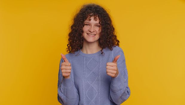 Like. Pretty curly haired woman in sweater raises thumbs up agrees with something or gives positive reply recommends advertisement likes good. Young girl isolated alone on yellow studio background