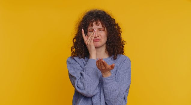 Upset disappointed young curly haired woman wipes tears and cries from despair, being sad because of unfair things. Teen girl in depression, despair, loss isolated alone on yellow studio background