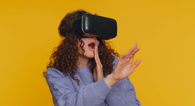 Young woman using headset helmet to play simulation game app online. Watching virtual reality 3D 360 video content. Girl in VR goggles isolated on studio yellow background. Futuristic technology