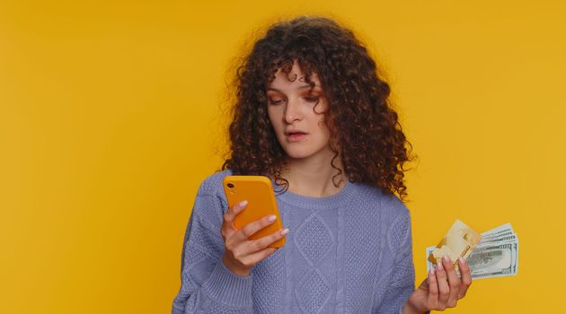 Sincere curly haired woman customer using credit bank card and smartphone while transferring money, purchases online shopping, payment. Finance and internet. Young girl isolated on yellow background