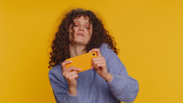 Worried funny addicted woman enthusiastically playing racing video games on mobile phone. Young curly haired girl using smartphone gadget app with drive simulator isolated on yellow studio background