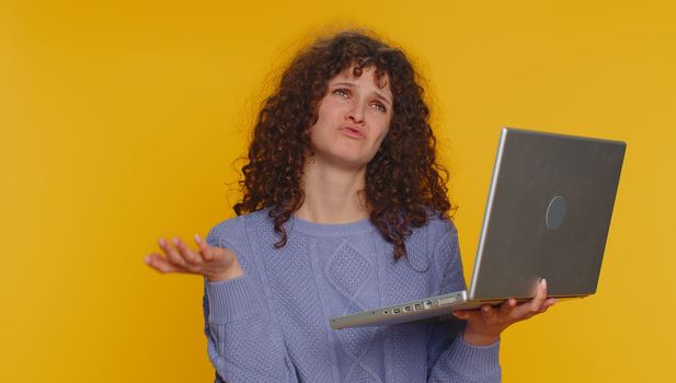 Young curly haired woman use laptop typing browsing working, loses becoming surprised sudden lottery results, bad news, fortune loss, fail, bankruptcy. Teen girl isolated on yellow studio background