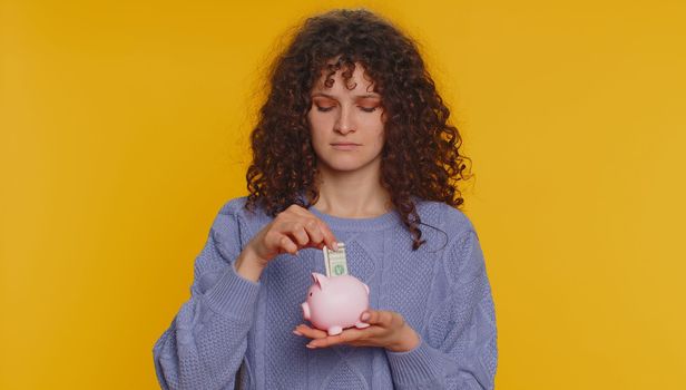 Poor young curly haired woman insufficient amount of money, holding piggybank and one dollar banknote. Financial crisis. Bankruptcy. Poverty and destitution. Teen girl on yellow studio background