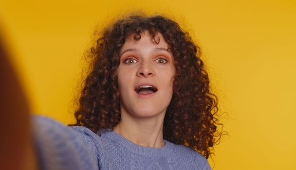 Oh my God, Wow. POV selfie portrait of excited amazed winner woman raising hands in surprise looking at camera with big eyes, shocked by sudden victory. Sincere curly haired girl on yellow background