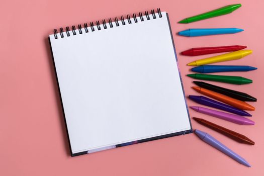 Open sketchbook with white empty blank. Colorful wax crayons on the pastel pink background. Flat lay composition