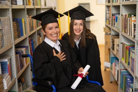 Happy young woman and woman in wheelchair in graduate gown with diploma in hands in library. Inclusive education