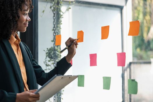 Attractive concentrated business lady in creating to-do list using multi coloured post-it sticky notes attaching them to transparent wall standing behind glass view, be more productive concept..