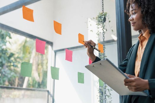 Attractive concentrated business lady in creating to-do list using multi coloured post-it sticky notes attaching them to transparent wall standing behind glass view, be more productive concept..