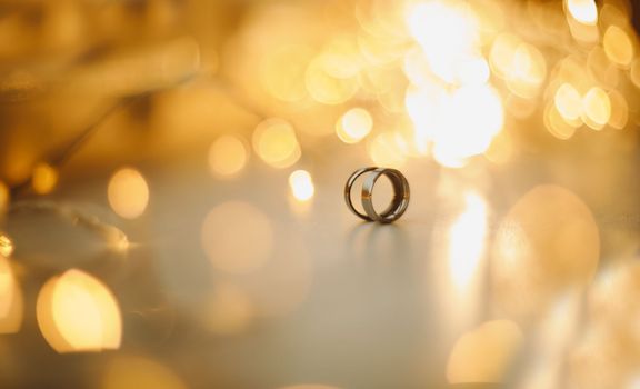 Wedding rings on blured bokeh glowing golden background. Symbol of love and romance on a textured glitter background with copy space for your greeting or congratulations.