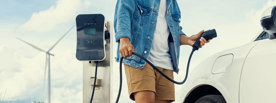 Progressive man with his electric car, EV car recharging energy from charging station on green field with wind turbine as concept of future sustainable energy. Electric vehicle with energy generator.