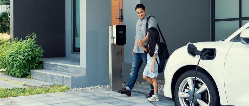 Progressive father and daughter plugs EV charger from home charging station to electric vehicle. Future eco-friendly car with EV cars powered by renewable source of clean energy.