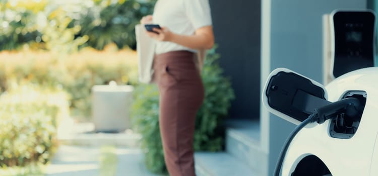 Focus image of electric vehicle recharging battery at home charging station with blurred woman walking in the background. Progressive concept of green energy technology applied in daily lifestyle.