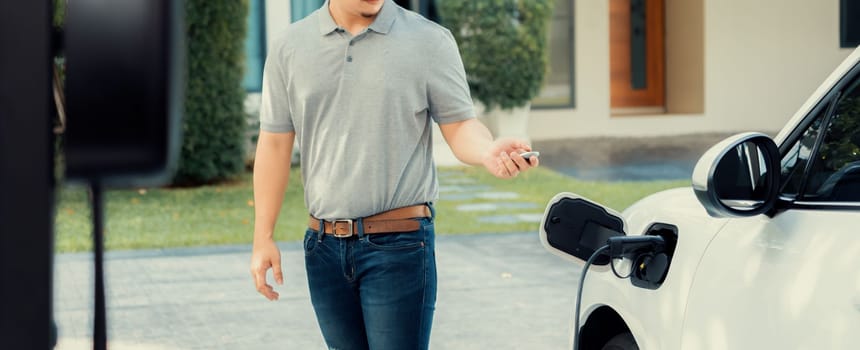 Progressive asian man install cable plug to his electric car with home charging station in the backyard. Concept use of electric vehicles in a progressive lifestyle contributes to clean environment.