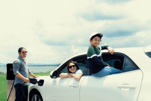 Concept of progressive happy family enjoying their time at green field and lake with electric vehicle. Electric vehicle driven by clean renewable from eco-friendly power sauce.