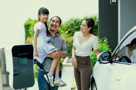 Progressive young parent teach daughter how to recharge or refuel EV car at home charging station. Green and clean energy from electric vehicle for healthy environment. Eco power from renewable source
