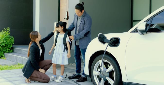 Progressive young parents and daughter with electric vehicle and home charging station. Green and clean energy from electric vehicles for healthy environment. Eco power from renewable source at home.